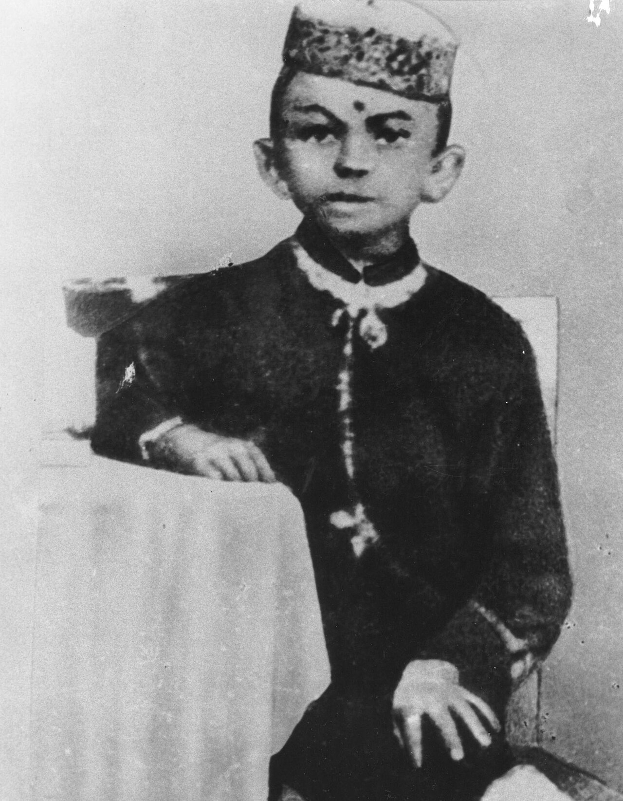 Black and white studio photograph of a young boy in a cap and a dark embroidered coat, sitting beside a draped table
