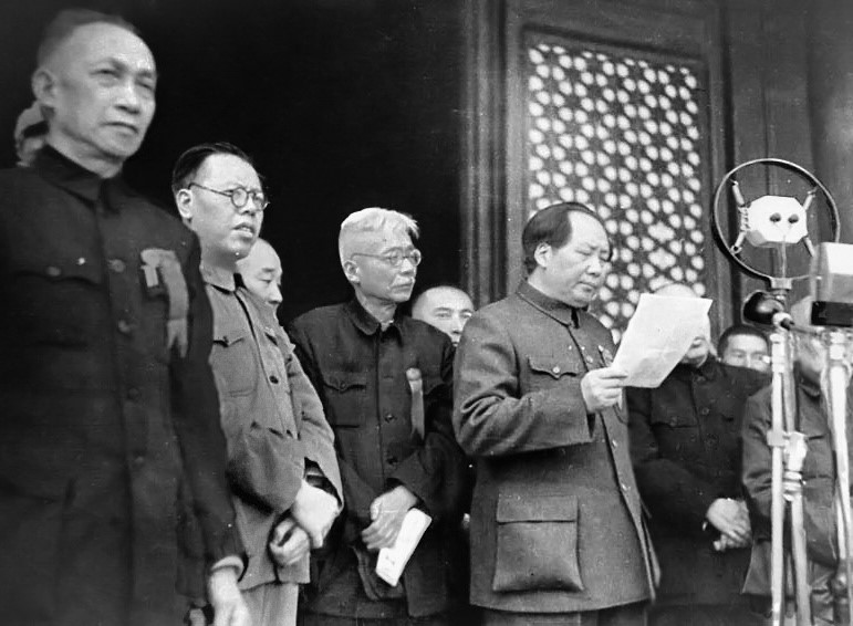 Black-and-white photograph of Mao Zedong reading from a sheet of paper at a microphone, with other leaders standing beside him on the Tiananmen Gate