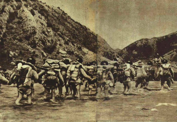 Black-and-white photograph of soldiers carrying large packs and rifles wading across a shallow river between rocky hills