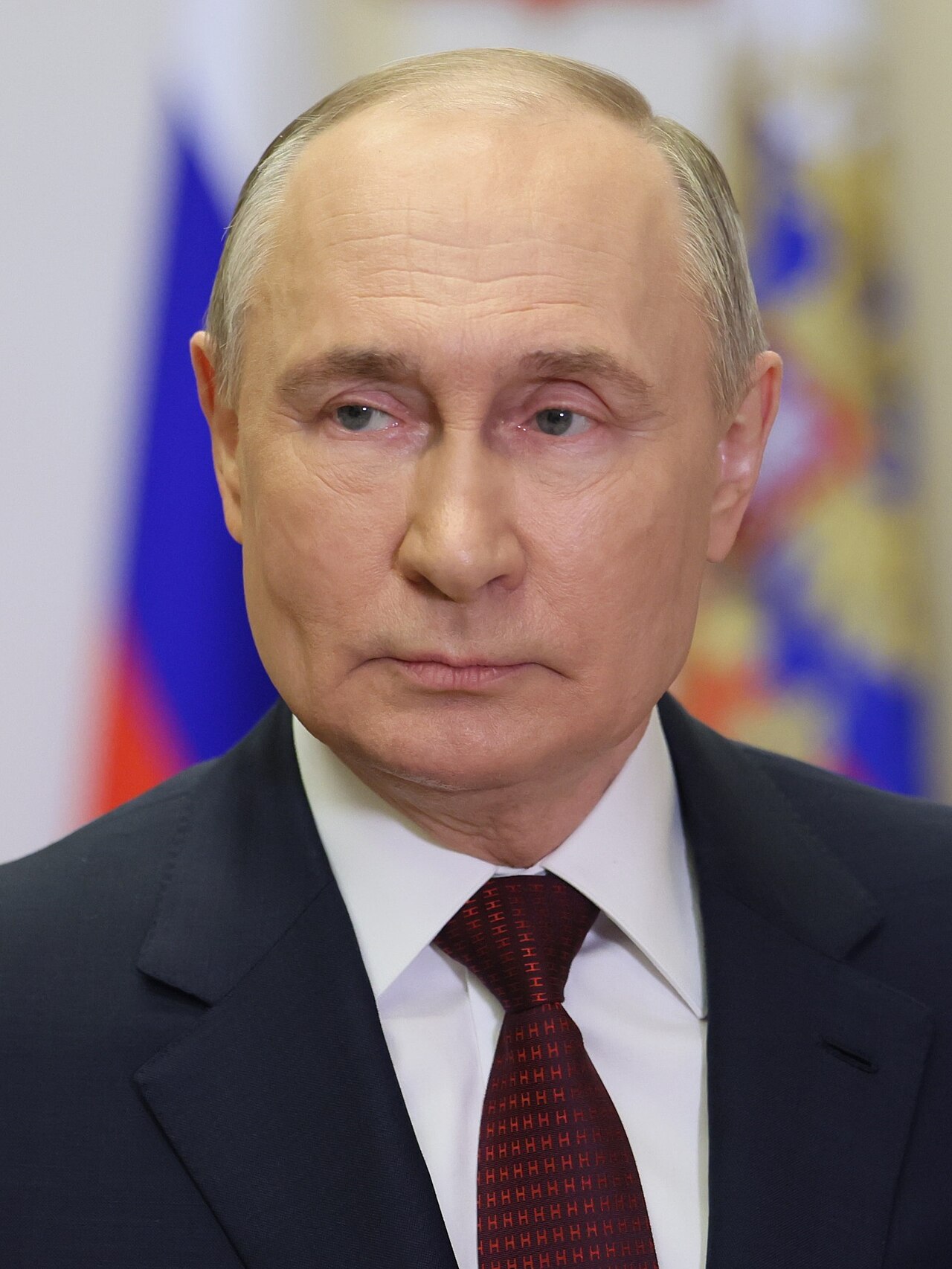 Head-and-shoulders portrait of Vladimir Putin in a dark suit and red tie, with a Russian flag behind him