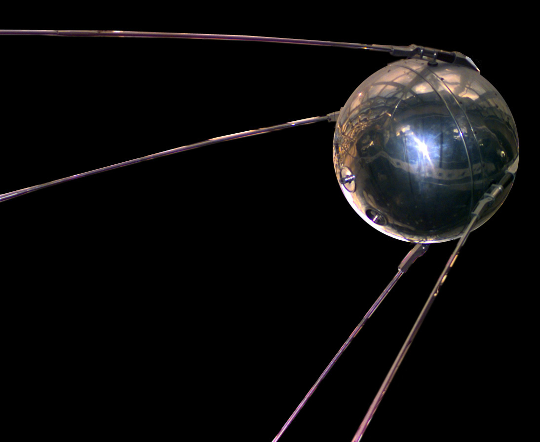 A metal sphere with four long, thin antennas trailing behind it, displayed against a dark background