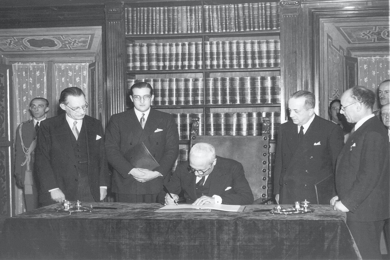 Black-and-white photograph of an older man in glasses signing a document at a draped table, with four men in suits standing around him in a book-lined room