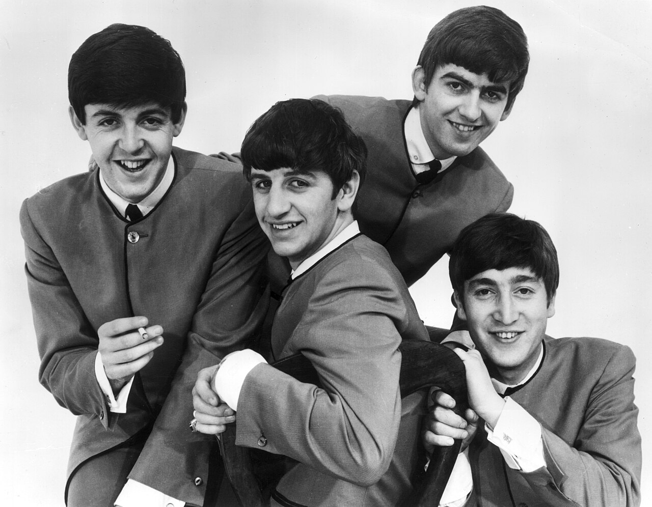 Black-and-white publicity photo of the four Beatles in matching suits, smiling, with Ringo Starr seated at the front
