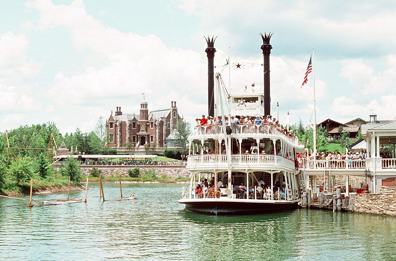 A white, double-decked riverboat crowded with passengers docks on a green river, with a brick mansion on a hill in the background