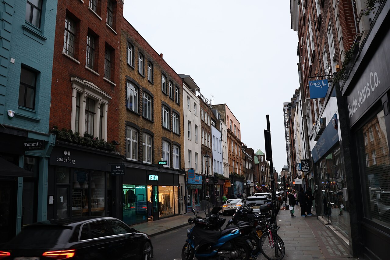 A narrow London street lined with brick buildings, shopfronts, parked motorcycles, and pedestrians on the sidewalk