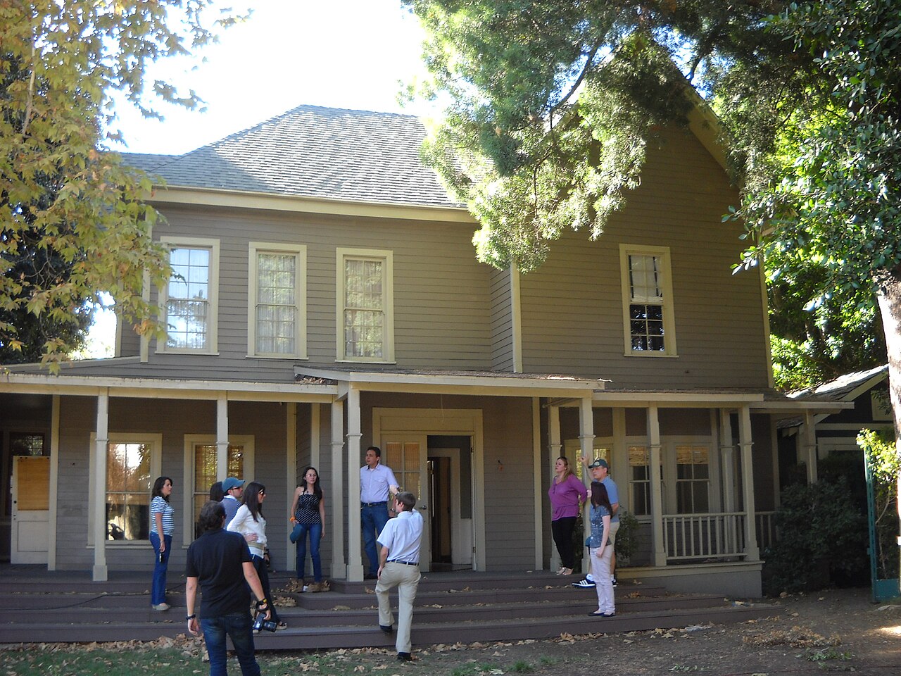 A two-story gray clapboard house with a wide front porch, shaded by trees, with a group of visitors standing on the steps