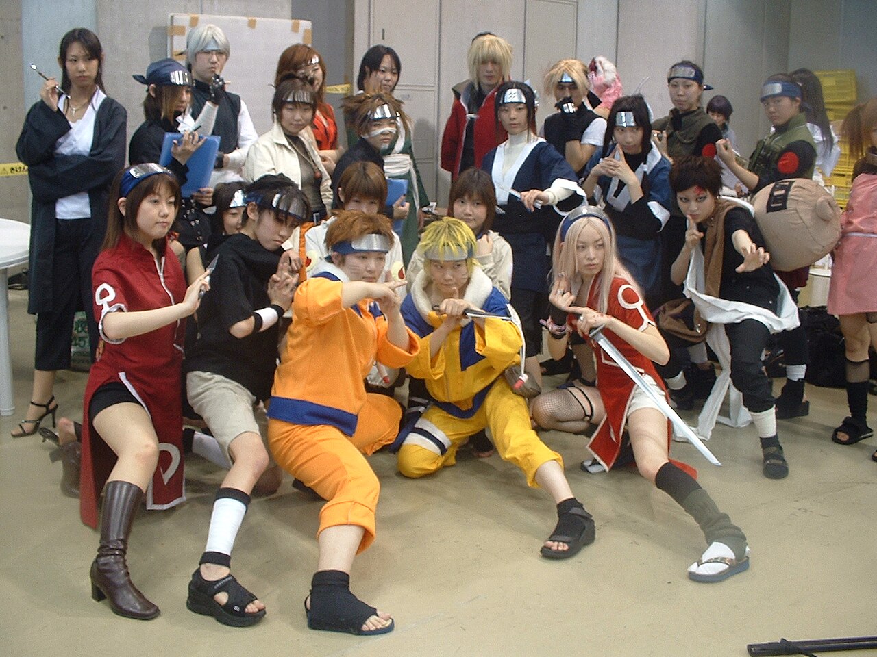 A large group of young cosplayers in ninja costumes and metal forehead protectors posing together, with two in orange and yellow outfits in the front row