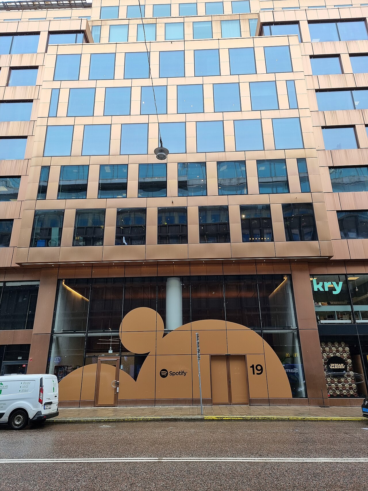 Street view of a tall office building with copper-colored panels and a large Spotify sign across its ground-floor windows