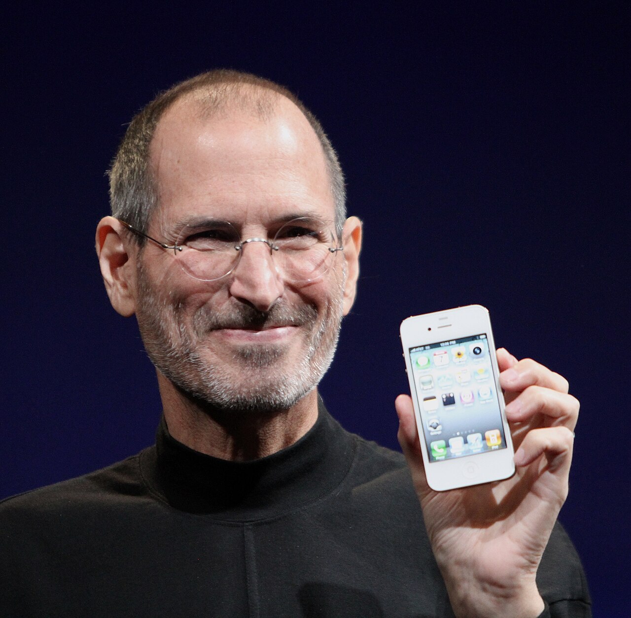Steve Jobs, smiling, in glasses and a black mock turtleneck, holding up a white iPhone 4 against a dark blue background