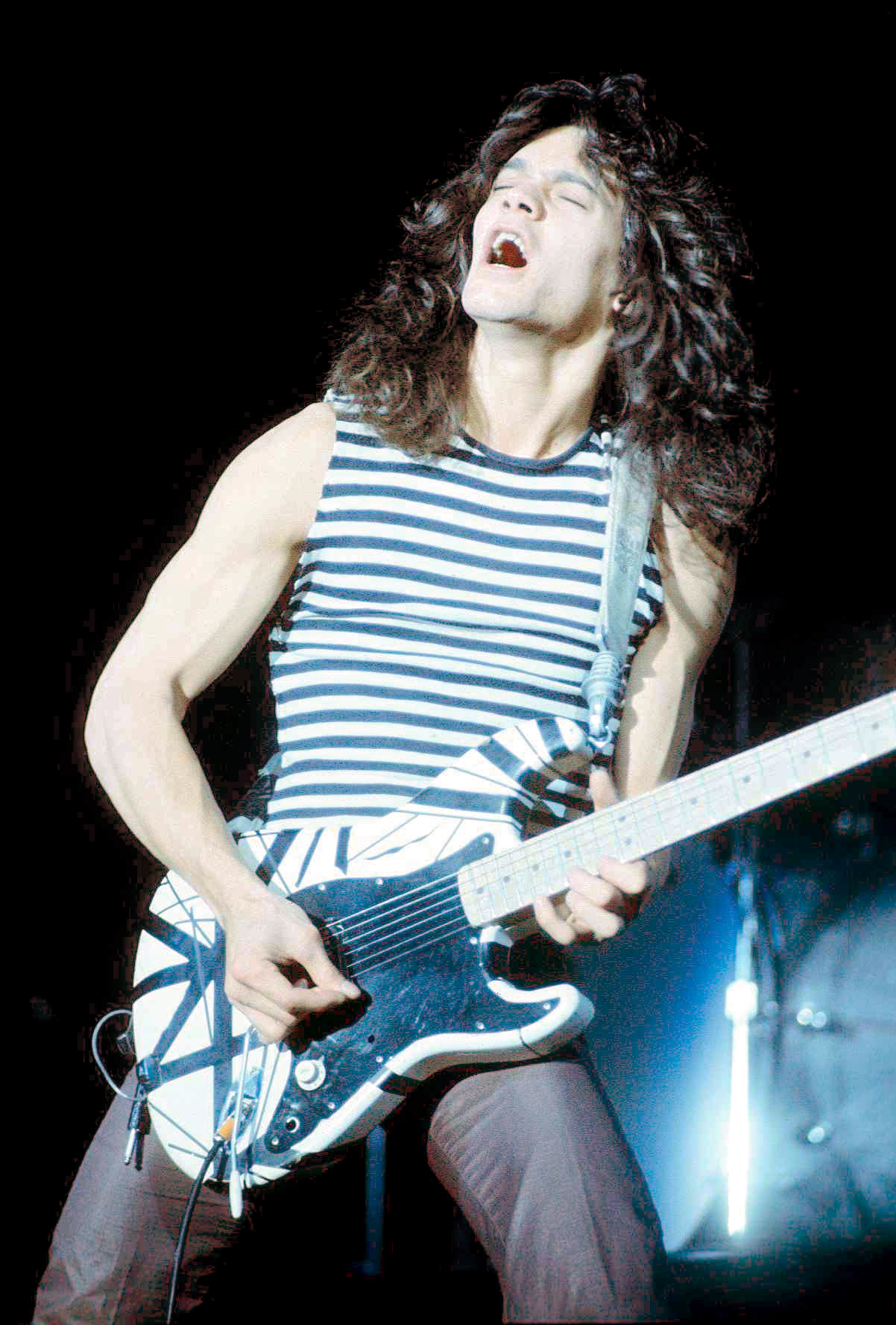 A young Eddie Van Halen, eyes closed, playing a black-and-white striped electric guitar on stage