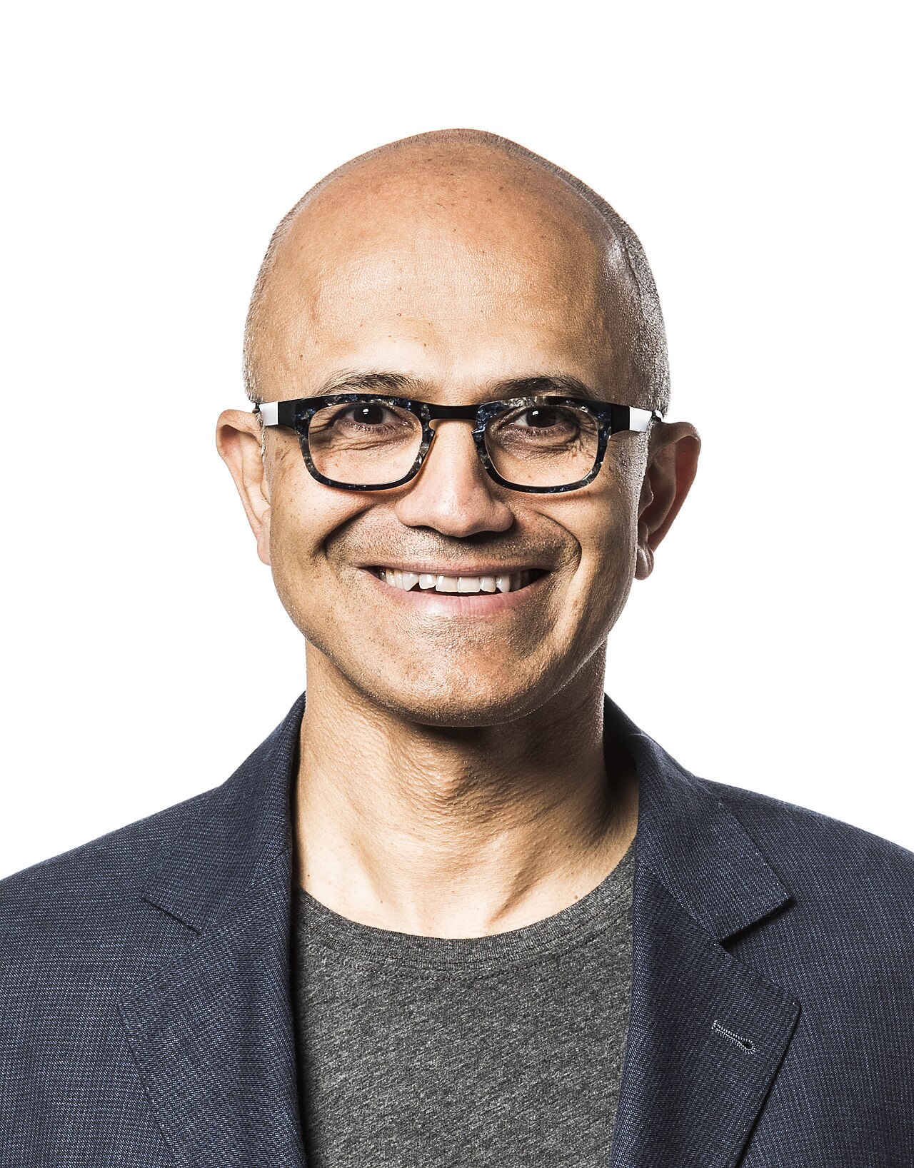 Portrait of Satya Nadella, a smiling bald man with dark-framed glasses, wearing a navy blazer over a gray T-shirt