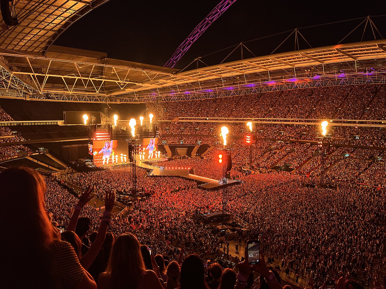 A packed stadium at night seen from the upper stands, with flames shooting from towers around the stage and Taylor Swift shown on giant screens